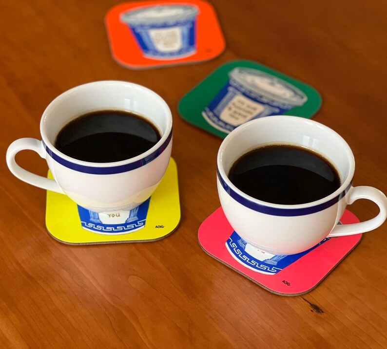 May include: Two white coffee mugs with black coffee on yellow and red coasters with a blue and white design. The coasters have the text "YOU" and "ASG" on them.