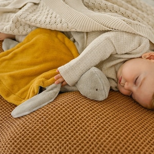 Può includere: Un morbido peluche con testa di coniglio grigio e lunghe orecchie attaccato a una coperta quadrata color giallo senape. Il giocattolo è realizzato in un tessuto vellutato e si trova accanto a un bambino che dorme sotto una coperta in maglia crema.