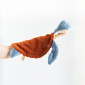 Puede incluir: Un juguete de peluche de tortuga marina con un caparazón texturizado de color naranja óxido y extremidades y cabeza de color azul claro. El juguete tiene una pequeña etiqueta de tela color canela con el texto "nuna" unido al lado del caparazón.