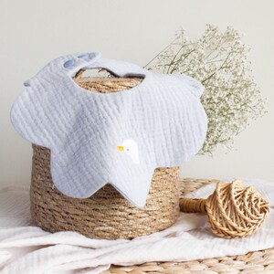 May include: A light blue baby bib with a scalloped edge and a white goose graphic. The bib is made of a soft, textured fabric and has two blue snap closures. The bib is resting on a woven basket.