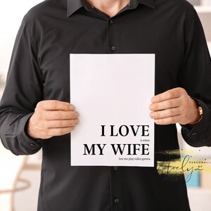 May include: A white sign with black text that reads "I love my wife, it when lets me play video games." The sign is held by a person wearing a black shirt.