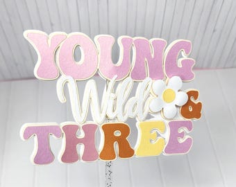 May include: A colorful cake topper with the words "Young Wild & Three" in pink, white, orange, and yellow. The topper has a glittery stick for insertion into a cake. The design includes a daisy and a brown ampersand.