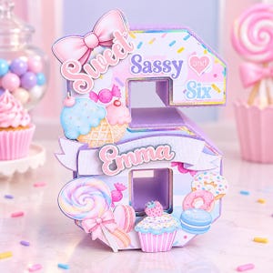 May include: A three-dimensional number six decoration in pastel colors, featuring the text Sweet Sassy and Six and the name Emma. The surface is decorated with illustrations of ice cream cones, lollipops, donuts, cupcakes, and a pink bow.