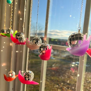 May include: A collection of disco ball necklaces hanging in front of a window. Each necklace features a small, mirrored disco ball pendant resting on a colorful, curved base. The bases are in shades of pink, orange, green, and purple.