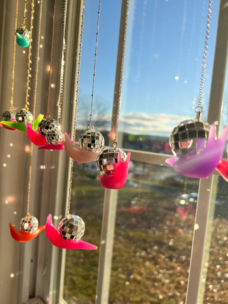 May include: A collection of disco ball necklaces hanging in front of a window. Each necklace features a small, mirrored disco ball pendant resting on a colorful, curved base. The bases are in shades of pink, orange, green, and purple.