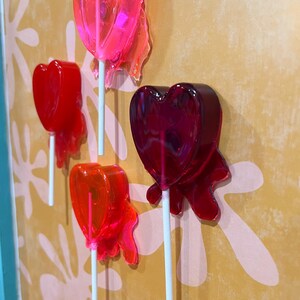 May include: Four heart-shaped lollipops in shades of red and pink are mounted on a wall. The lollipops are on white sticks, with a translucent, glossy appearance. The wall has a yellow background with a floral pattern.