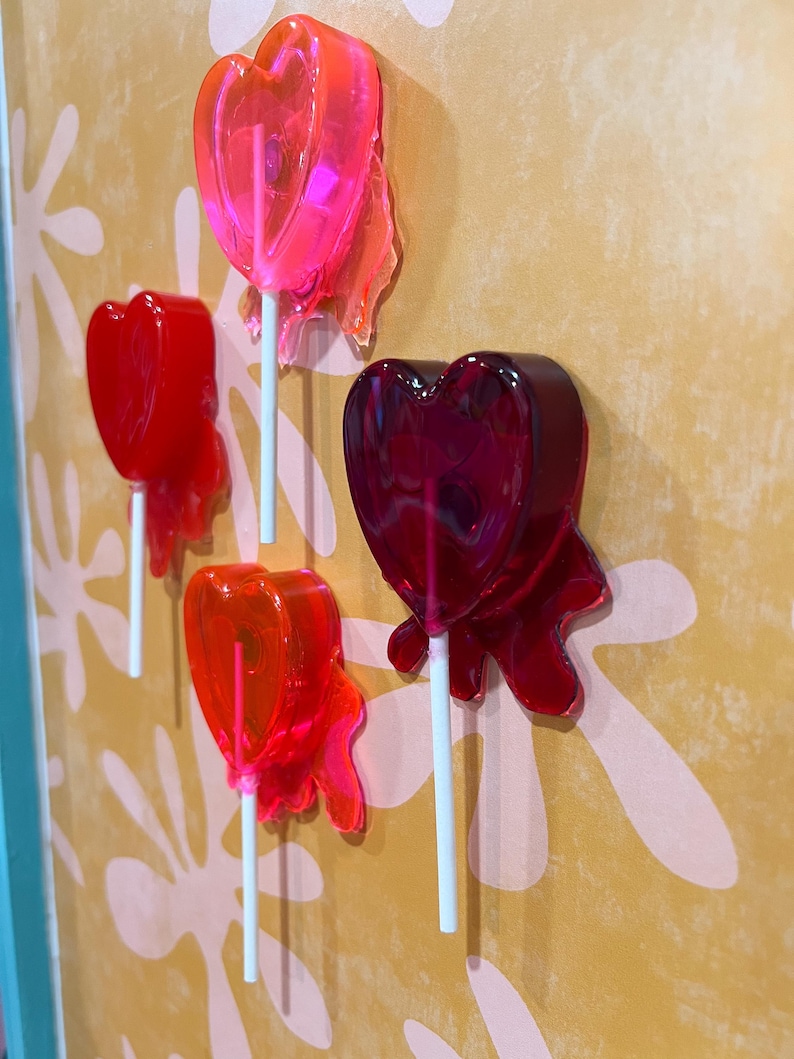 May include: Four heart-shaped lollipops in shades of red and pink are mounted on a wall. The lollipops are on white sticks, with a translucent, glossy appearance. The wall has a yellow background with a floral pattern.