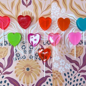 May include: A selection of heart-shaped lollipops in a range of colours, such as red, orange, green, pink, and blue, set against a floral patterned backdrop. Each lollipop is mounted on a white stick, creating a bright and cheerful arrangement.