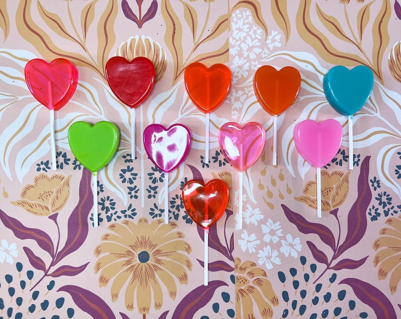 May include: A selection of heart-shaped lollipops in a range of colours, such as red, orange, green, pink, and blue, set against a floral patterned backdrop. Each lollipop is mounted on a white stick, creating a bright and cheerful arrangement.
