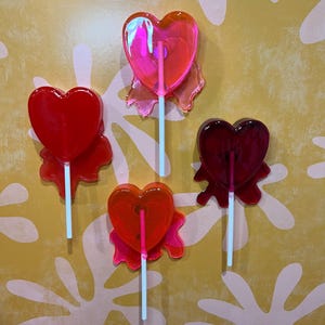 May include: Four heart-shaped lollipops in shades of red and pink, each with a white stick. The lollipops have a translucent appearance and are arranged against a yellow background with a white floral pattern.