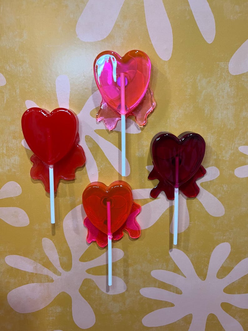 May include: Four heart-shaped lollipops in shades of red and pink, each with a white stick. The lollipops have a translucent appearance and are arranged against a yellow background with a white floral pattern.