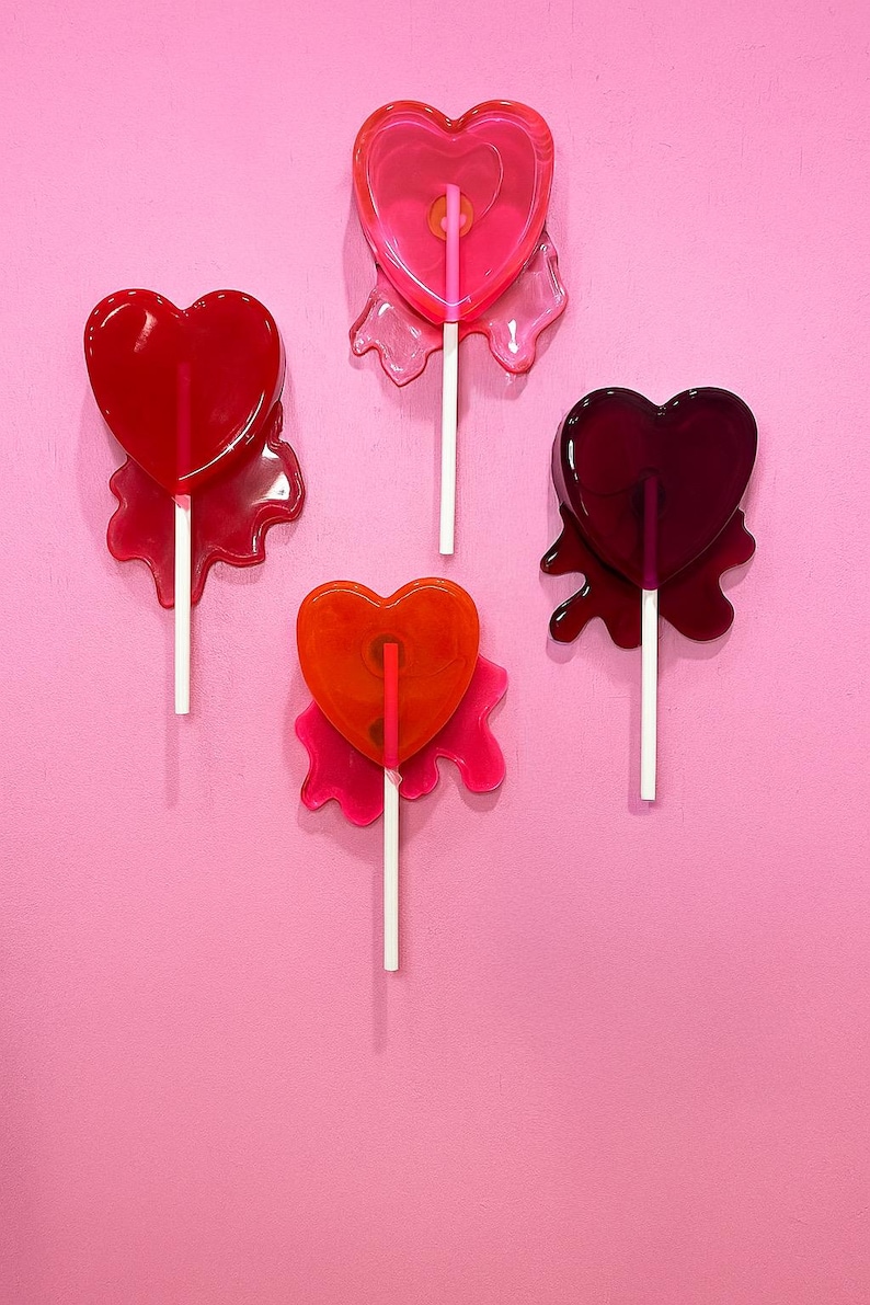 May include: Four heart-shaped lollipops in shades of red, orange, and pink, each on a white stick, against a pink background. The lollipops have a melted, dripping effect at the base, creating a playful and colorful design.