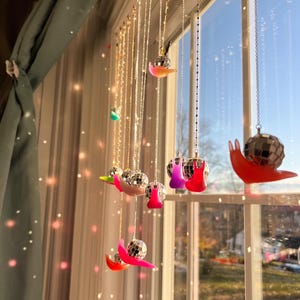May include: A window display featuring multiple strands of decorative chains, each adorned with a miniature disco ball and a colorful, snail-shaped accent. The accents are in shades of red, pink, purple, and green. The window is framed by a curtain.
