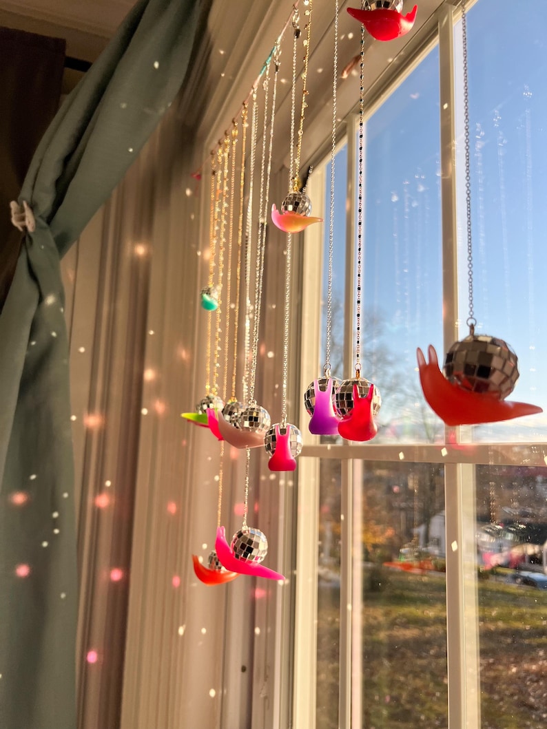 May include: A window display featuring multiple strands of decorative chains, each adorned with a miniature disco ball and a colorful, snail-shaped accent. The accents are in shades of red, pink, purple, and green. The window is framed by a curtain.