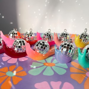 May include: A collection of colorful snail figurines, each topped with a miniature disco ball. The snails are in shades of pink, red, orange, yellow, green, and purple, set against a floral patterned background.