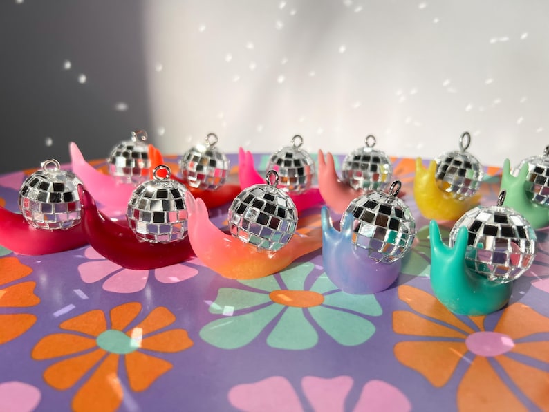 May include: A collection of colorful snail figurines, each topped with a miniature disco ball. The snails are in shades of pink, red, orange, yellow, green, and purple, set against a floral patterned background.