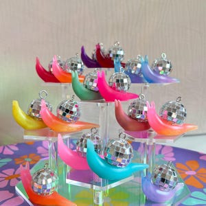 May include: A display of colorful snail ornaments, each with a small disco ball shell. The snails are in various shades of pink, orange, green, blue, purple, and yellow. They are arranged on clear acrylic stands.