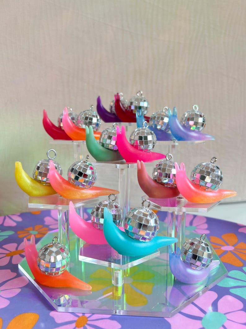 May include: A display of colorful snail ornaments, each with a small disco ball shell. The snails are in various shades of pink, orange, green, blue, purple, and yellow. They are arranged on clear acrylic stands.