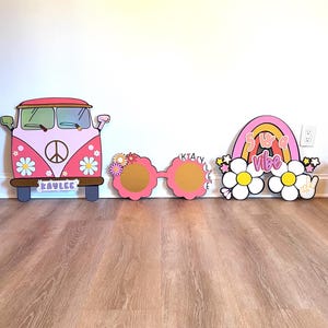 May include: Pink and white wooden cutouts with a retro theme. Includes a pink van with a peace sign, large pink sunglasses, and a rainbow with the text "5 a vibe". The van has the name "Kaylee" on it.