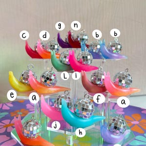 May include: A collection of colorful snail ornaments, each with a disco ball shell. The snails are in various shades including pink, orange, blue, and purple. They are displayed on a clear, multi-tiered stand.