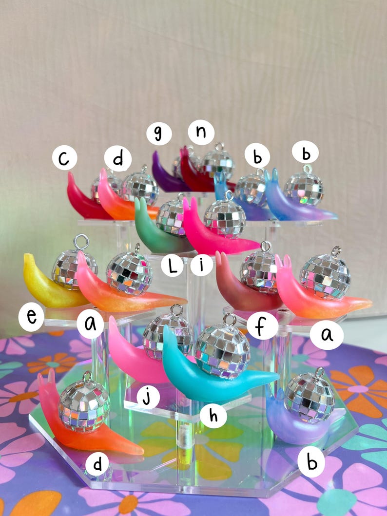 May include: A collection of colorful snail ornaments, each with a disco ball shell. The snails are in various shades including pink, orange, blue, and purple. They are displayed on a clear, multi-tiered stand.