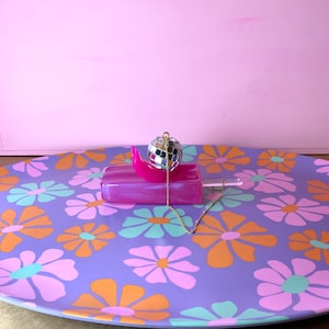 May include: A purple floral tray with orange, pink, and teal flowers. A pink popsicle-shaped object with a disco ball and gold chain necklace are on the tray. The background is a solid pink.