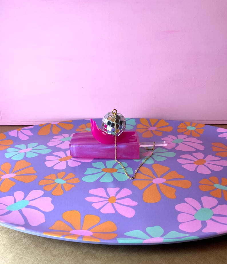May include: A purple floral tray with orange, pink, and teal flowers. A pink popsicle-shaped object with a disco ball and gold chain necklace are on the tray. The background is a solid pink.