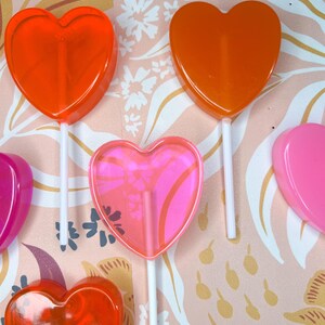 May include: A selection of heart-shaped lollipops in red, orange, and pink. The lollipops are on white sticks, with one featuring a floral design. The background has a pink and white floral pattern.