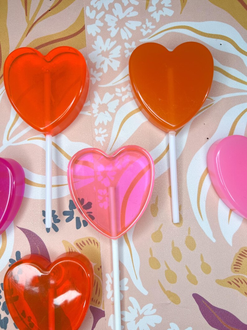 May include: A selection of heart-shaped lollipops in red, orange, and pink. The lollipops are on white sticks, with one featuring a floral design. The background has a pink and white floral pattern.