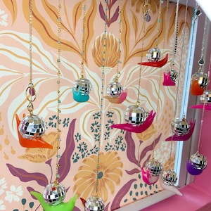 May include: A display of hanging disco ball pendants, each with a small, colorful snail-shaped base. The pendants are suspended from gold-colored chains against a floral wallpaper backdrop. The snail bases are in various colors, including orange, pink, green, and purple.