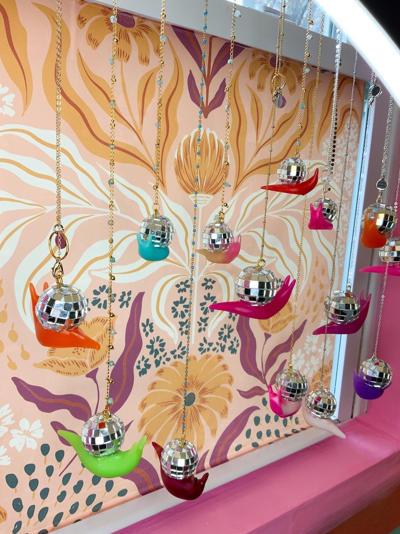 May include: A display of hanging disco ball pendants, each with a small, colorful snail-shaped base. The pendants are suspended from gold-colored chains against a floral wallpaper backdrop. The snail bases are in various colors, including orange, pink, green, and purple.