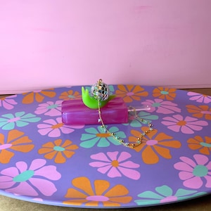 May include: A decorative purple tray with a floral pattern in orange, pink, and green. A green snail-shaped object holds a disco ball and a pink rectangular object. A gold chain with small beads is draped across the tray.