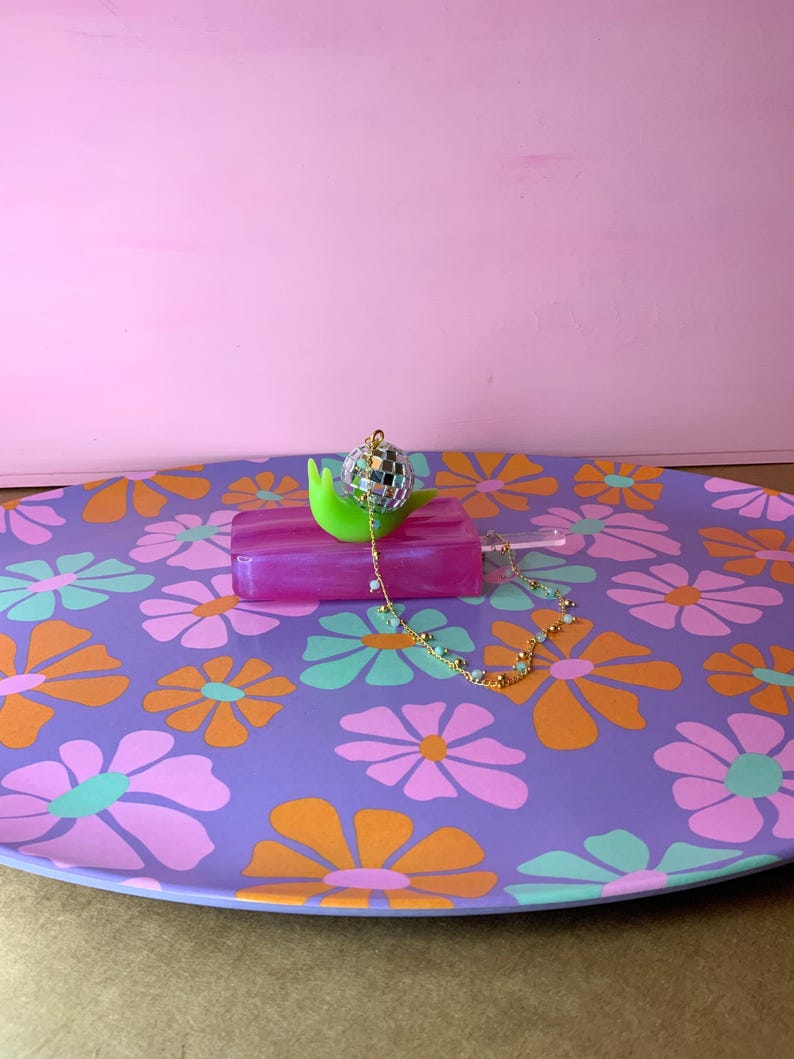 May include: A decorative purple tray with a floral pattern in orange, pink, and green. A green snail-shaped object holds a disco ball and a pink rectangular object. A gold chain with small beads is draped across the tray.