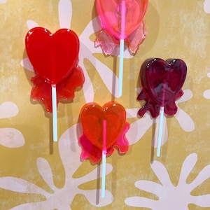 May include: Four heart-shaped lollipops in shades of red, orange, and pink. Each lollipop has a light blue stick and a decorative, translucent base. The lollipops are arranged against a yellow background with white floral patterns.