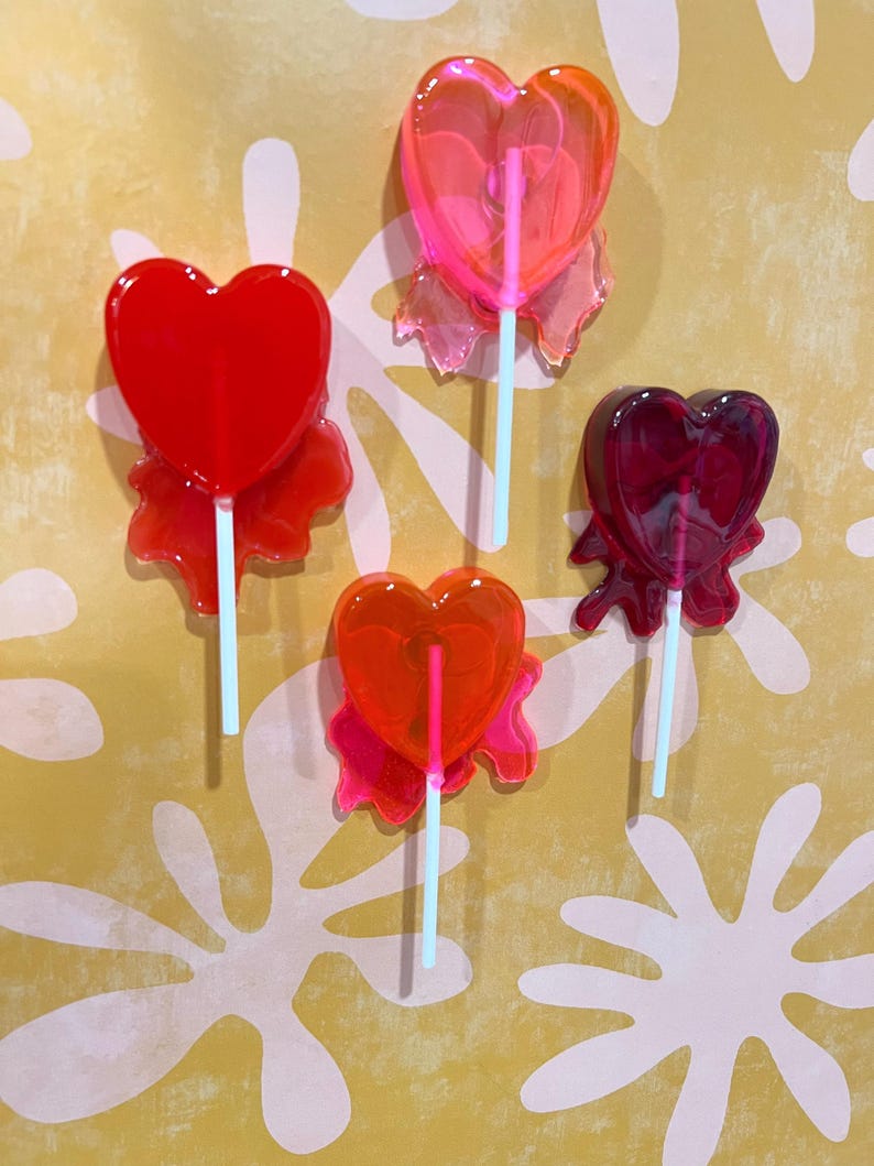 May include: Four heart-shaped lollipops in shades of red, orange, and pink. Each lollipop has a light blue stick and a decorative, translucent base. The lollipops are arranged against a yellow background with white floral patterns.