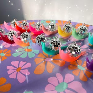 May include: A collection of colorful snail-shaped ornaments, each topped with a miniature disco ball. The snails are in various bright colors, including red, pink, yellow, green, orange, and purple, arranged on a floral patterned surface.