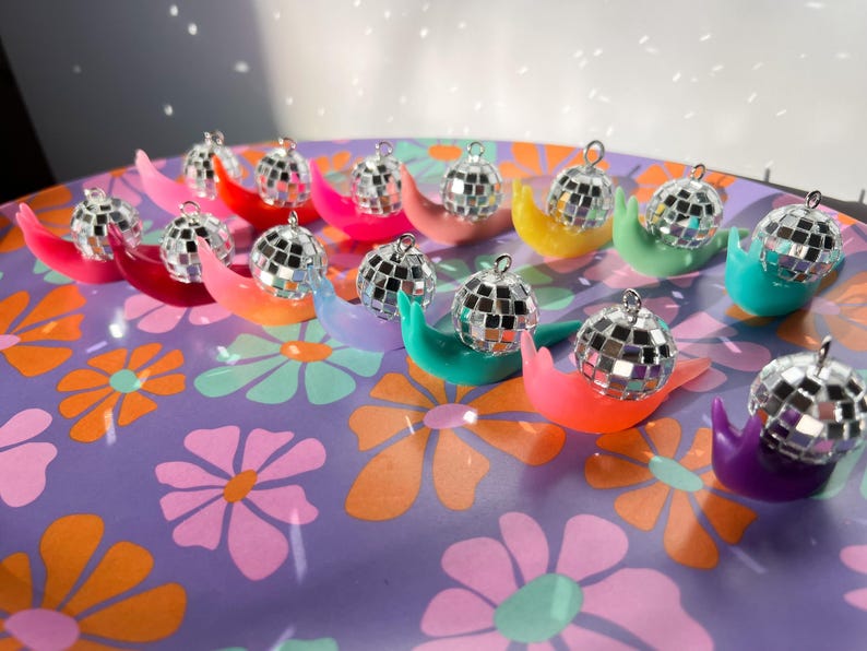 May include: A collection of colorful snail-shaped ornaments, each topped with a miniature disco ball. The snails are in various bright colors, including red, pink, yellow, green, orange, and purple, arranged on a floral patterned surface.