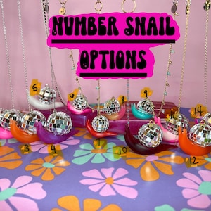 May include: A collection of colorful snail-shaped necklaces with disco ball pendants. The necklaces are displayed on a floral patterned surface. The sign above reads "NUMBER SNAIL OPTIONS". Each snail is a different color and numbered.