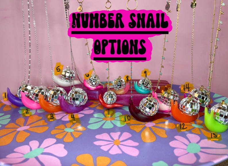 May include: A collection of colorful snail-shaped necklaces with disco ball pendants. The necklaces are displayed on a floral patterned surface. The sign above reads "NUMBER SNAIL OPTIONS". Each snail is a different color and numbered.