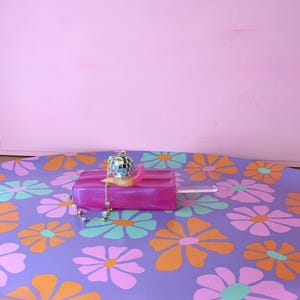 May include: A decorative purple tray with a floral pattern in orange, pink, and teal. A pink popsicle-shaped object with a disco ball charm and a gold chain sits on the tray. The background is a solid pink.