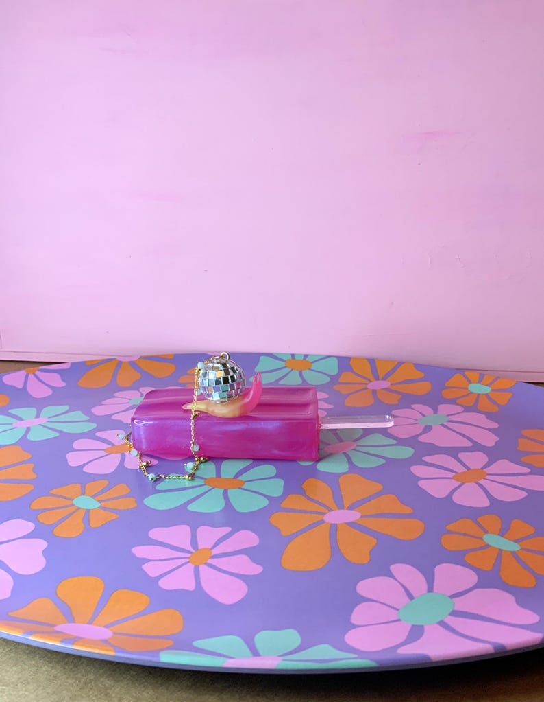 May include: A decorative purple tray with a floral pattern in orange, pink, and teal. A pink popsicle-shaped object with a disco ball charm and a gold chain sits on the tray. The background is a solid pink.