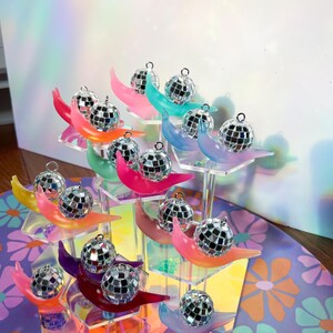 May include: A display of miniature disco ball ornaments on colorful crescent-shaped stands. The stands are arranged on a multi-tiered, clear acrylic display. The crescent stands are in various colors, including pink, blue, and yellow. The background is a floral pattern.