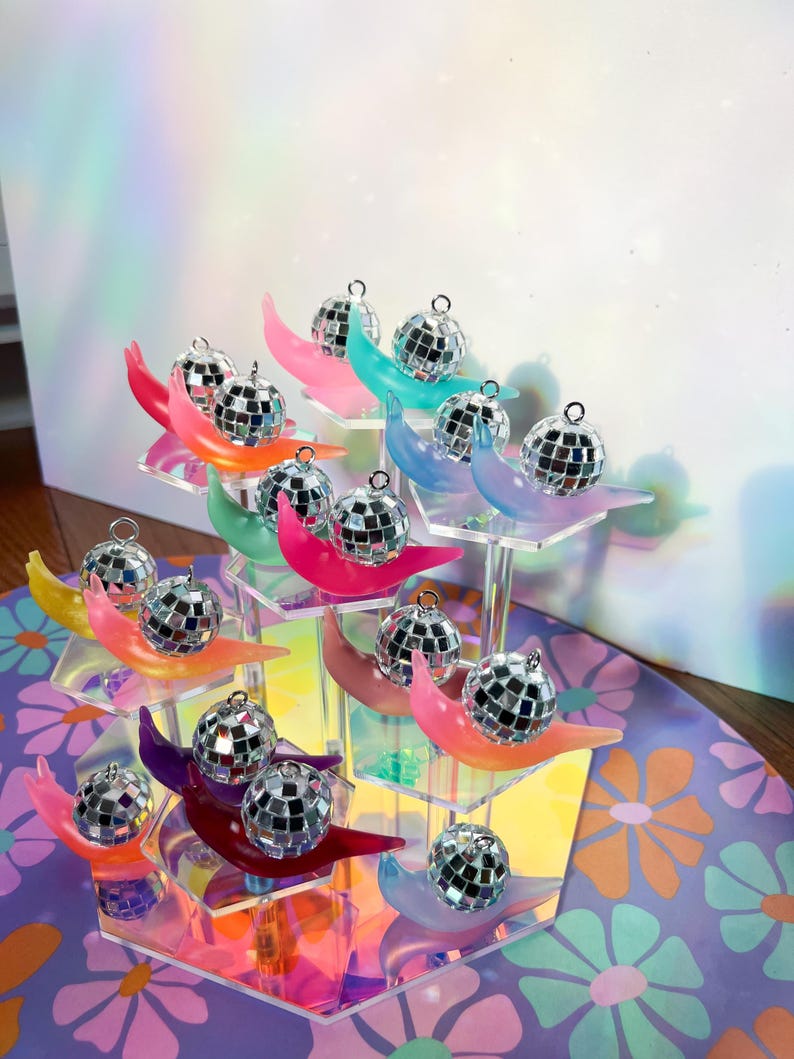 May include: A display of miniature disco ball ornaments on colorful crescent-shaped stands. The stands are arranged on a multi-tiered, clear acrylic display. The crescent stands are in various colors, including pink, blue, and yellow. The background is a floral pattern.