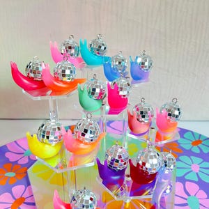 May include: A tiered display of colorful snail-shaped ornaments, each holding a small disco ball. The snails are in shades of pink, orange, yellow, green, blue, and purple. The display sits on a floral patterned surface.