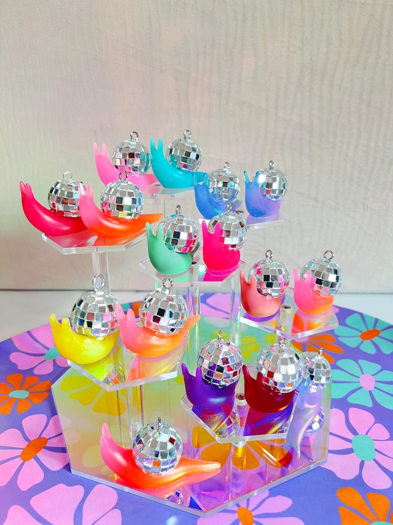 May include: A tiered display of colorful snail-shaped ornaments, each holding a small disco ball. The snails are in shades of pink, orange, yellow, green, blue, and purple. The display sits on a floral patterned surface.