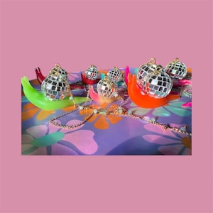 May include: A collection of small disco ball ornaments, each with a gold-colored loop, resting on colorful, curved holders. A gold-colored chain with small white beads is draped across the scene. The background is a patterned purple surface.