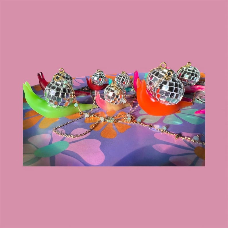 May include: A collection of small disco ball ornaments, each with a gold-colored loop, resting on colorful, curved holders. A gold-colored chain with small white beads is draped across the scene. The background is a patterned purple surface.