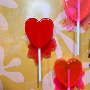 May include: Several heart-shaped lollipops in shades of red and pink are displayed against a yellow background with white floral patterns. Each lollipop has a white stick and a translucent appearance, with a glossy finish.