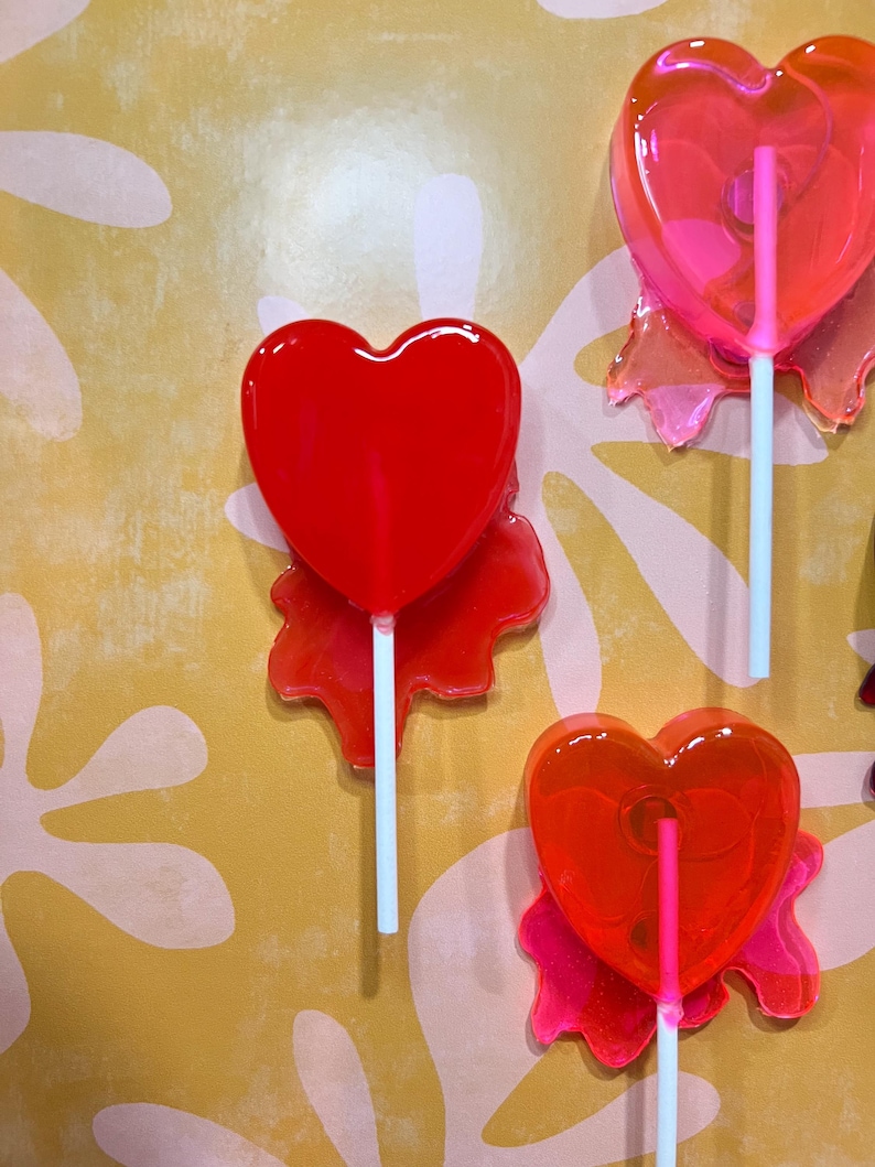 May include: Several heart-shaped lollipops in shades of red and pink are displayed against a yellow background with white floral patterns. Each lollipop has a white stick and a translucent appearance, with a glossy finish.