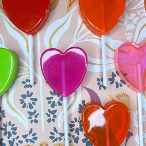 May include: A selection of heart-shaped lollipops in shades of red, orange, pink, and green, each on a white stick. The lollipops are displayed against a patterned backdrop with floral and leaf designs.